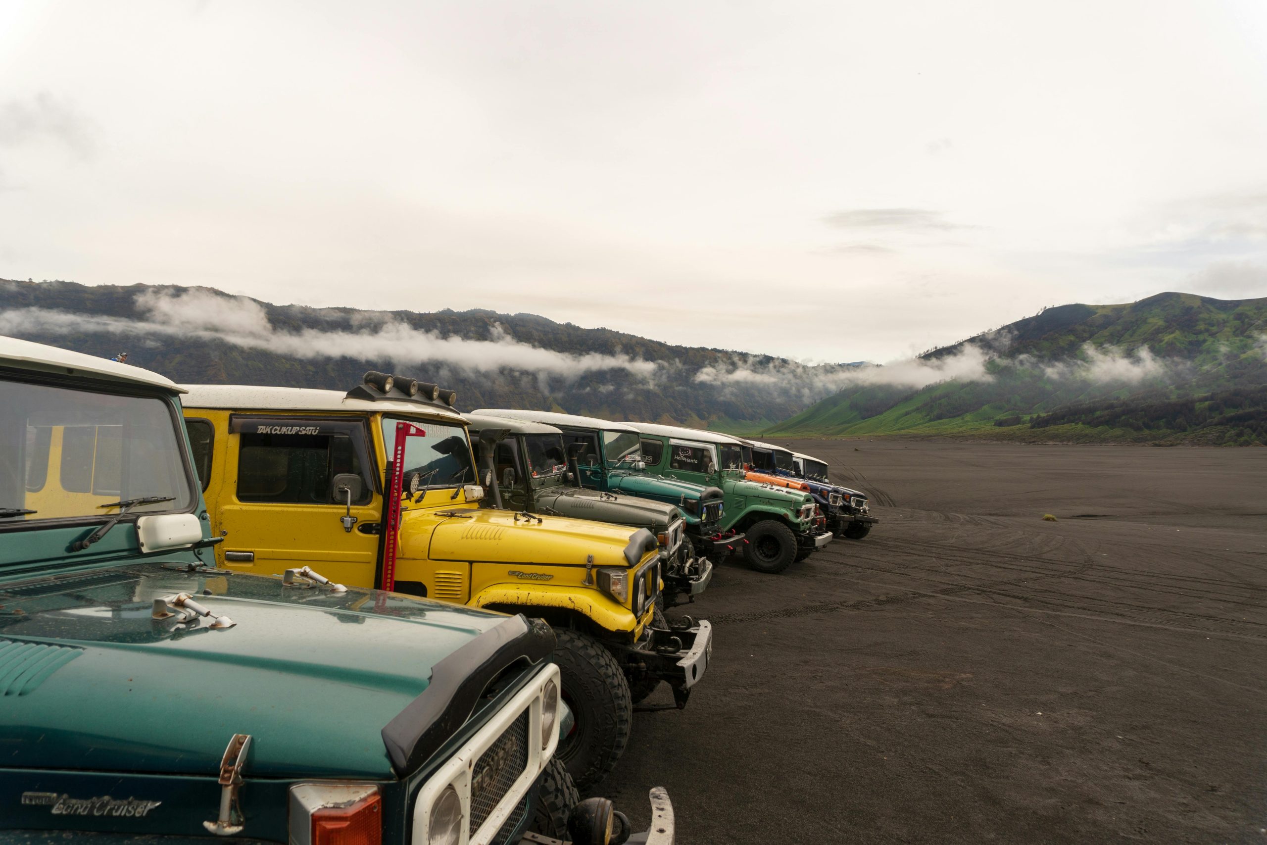 Classic offroad vehicles line up on volcanic terrain near mist-covered Mount Bromo.